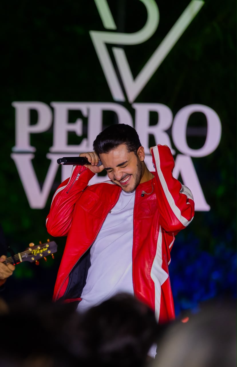 Pedro Vieira em show, usando jaqueta vermelha diante da logomarca